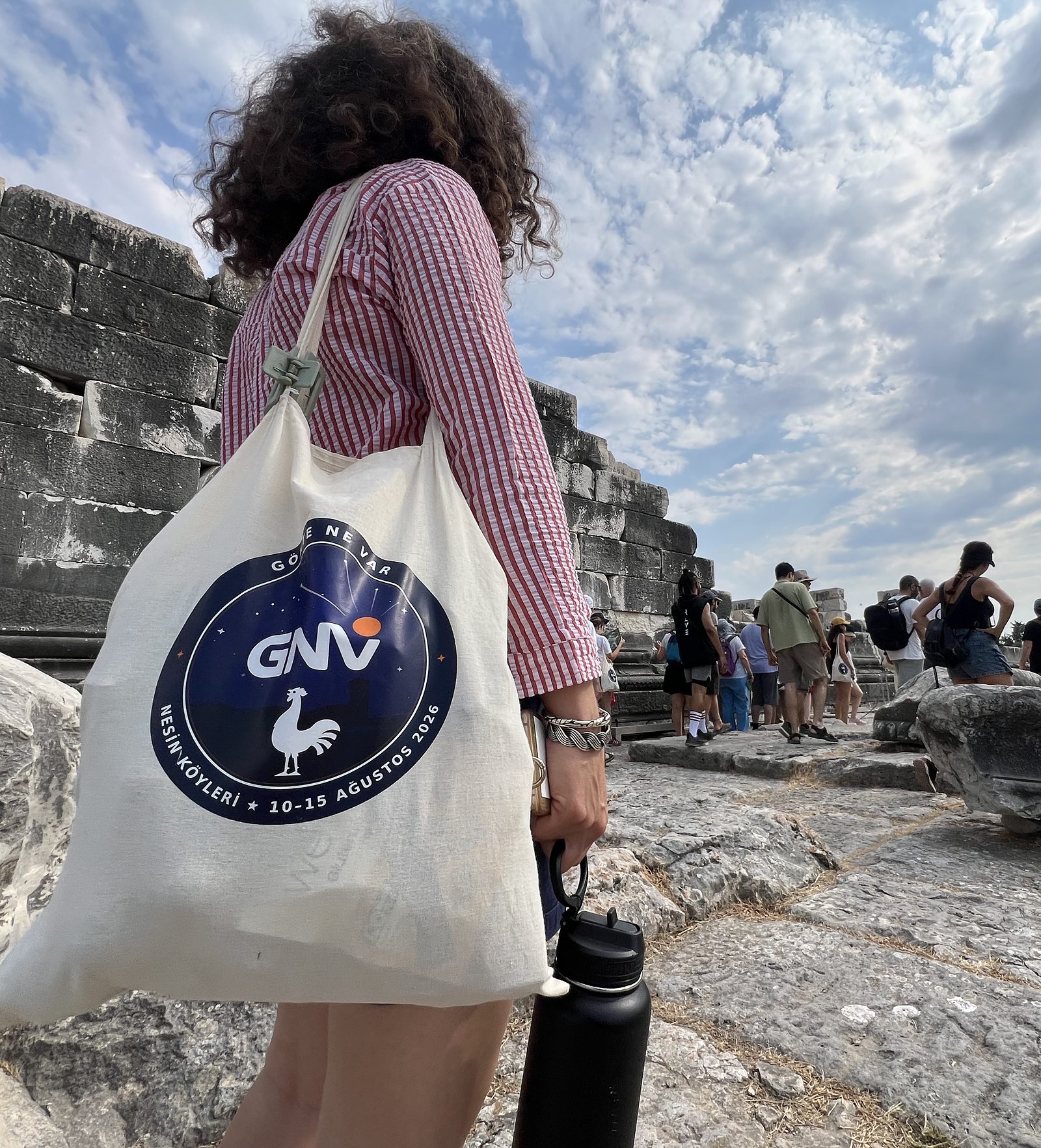 Didim’deki Apollon Tapınağı’nda, omzunda Gökte Ne Var çantasıyla kalıntılara doğru yürüyen bir katılımcı; ileride tapınağı gezen grup ve bulutlu gökyüzü